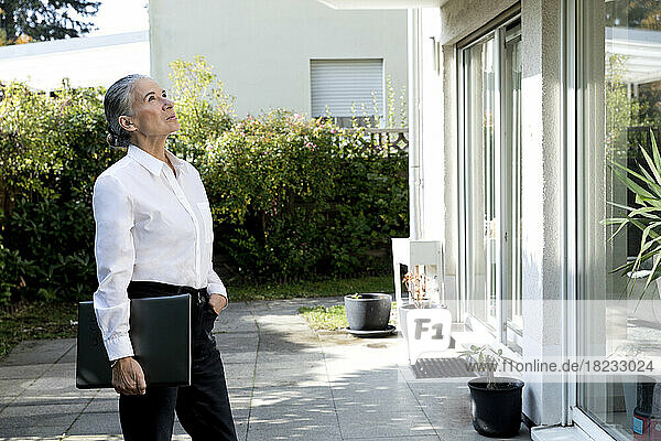 Ältere Frau hält Laptop  steht mit der Hand in der Tasche und blickt auf das Haus