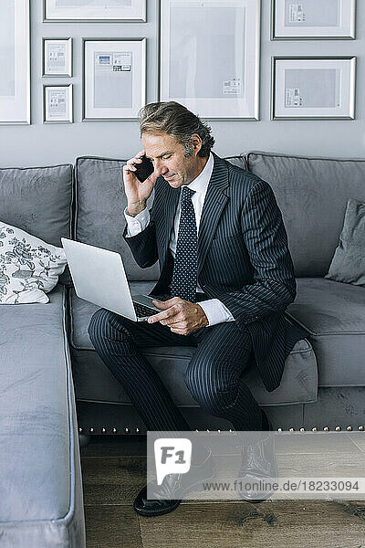 Mature businessman sitting with laptop and talking through smart phone at home