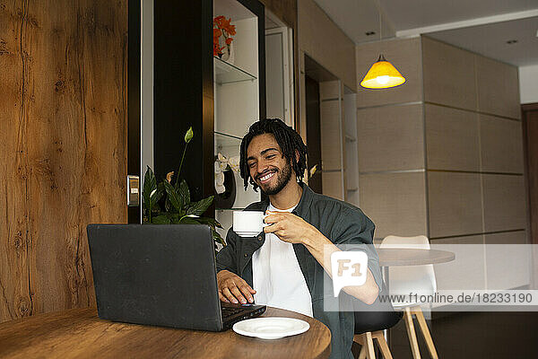 Glücklicher Freiberufler hält Teetasse in der Hand und schaut im Heimbüro auf den Laptop