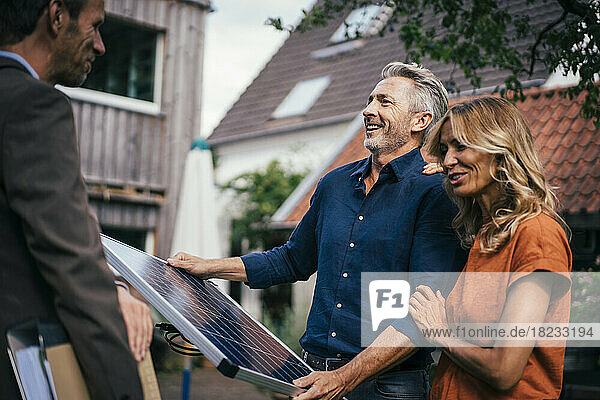 Glückliches Paar hält Solarpanel in der Hand und diskutiert mit Immobilienmakler im Hinterhof