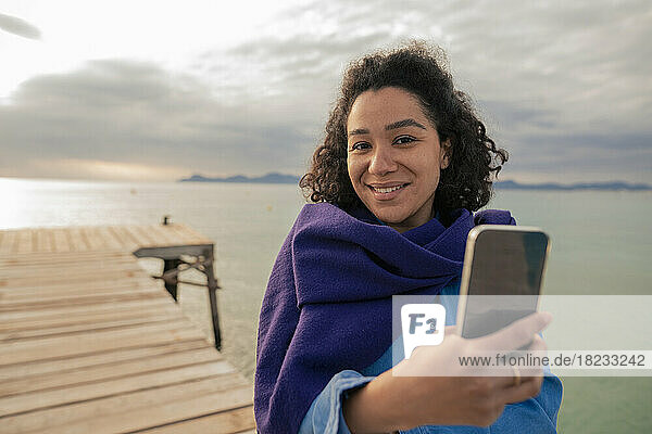 Woman with smart phone taking selfie on jetty by sea