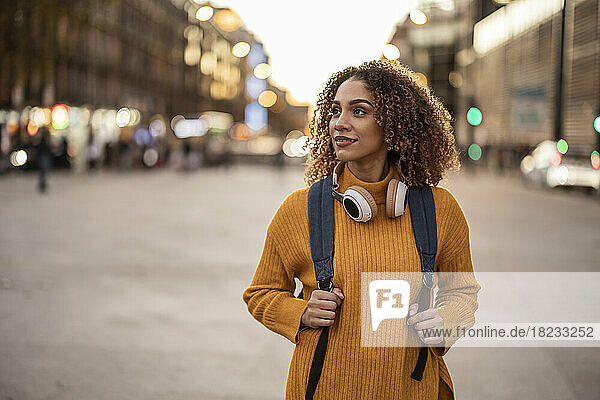 Young woman with backpack and wireless headphones walking on footpath at sunset