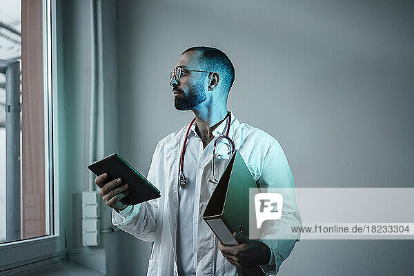 Thoughtful doctor holding tablet PC and file folder looking through window in hospital