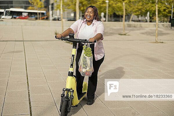 Glückliche Frau mit Elektroroller und Tüte Gemüse steht am Fußweg