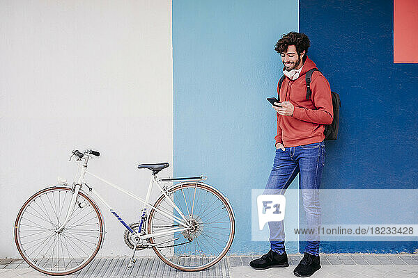 Smiling man using smart phone by bicycle near colorful wall