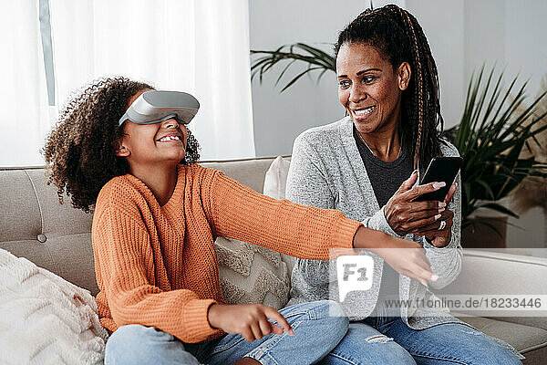 Smiling mother with daughter wearing virtual reality simulator on sofa at home