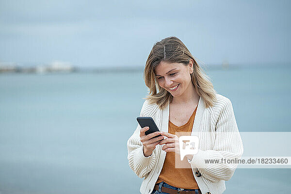 Smiling beautiful young woman text messaging on mobile phone at beach