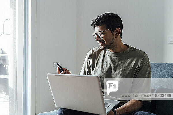 Smiling man using smart phone sitting with laptop on sofa