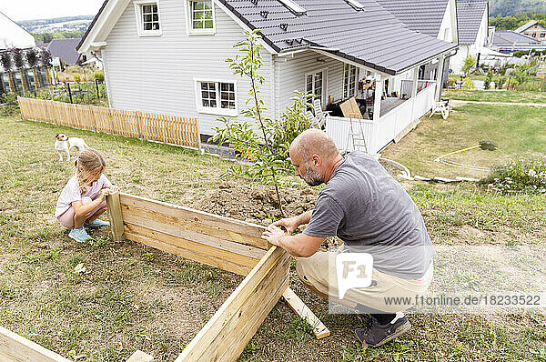 Father and daughter making raised bed crouching in back yard near house