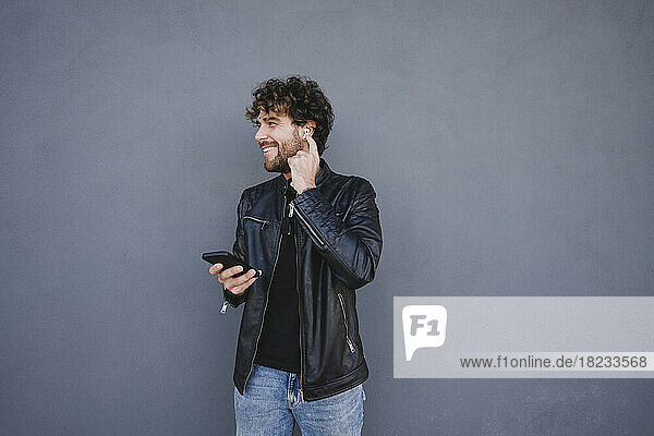 Glücklicher Mann hält sein Mobiltelefon in der Hand und stellt drahtlose In-Ear-Kopfhörer vor einer grauen Wand ein
