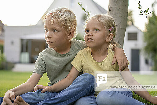 Bruder sitzt mit Arm um Schwester auf Gras im Garten