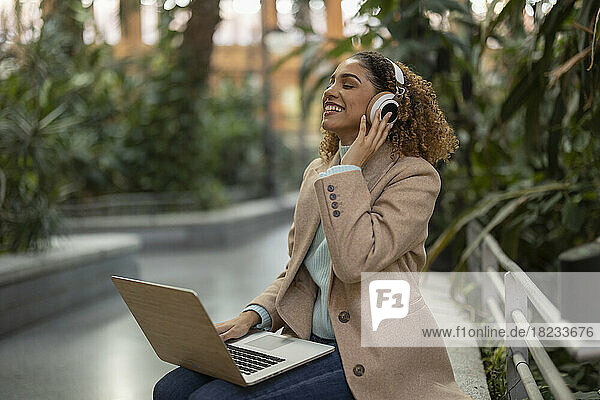 Happy businesswoman listening to music through wireless headphones sitting with laptop on bench