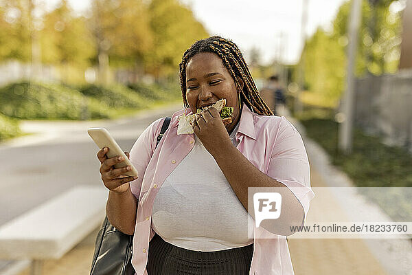Junge Frau benutzt Smartphone und isst Sandwich  das am Fußweg steht