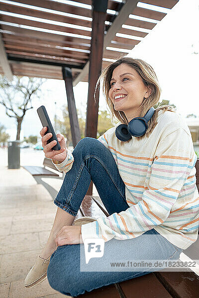 Happy young woman sitting on bench with smart phone and wireless headphones