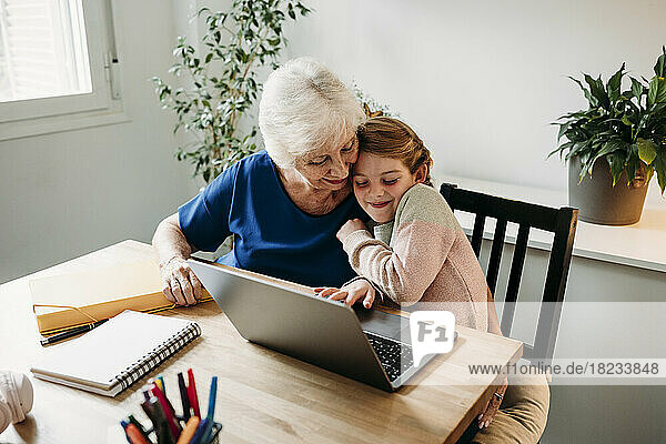 Smiling girl with laptop on table embracing grandmother at home