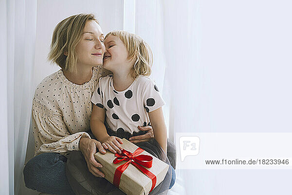 Mother and daughter with Christmas present sitting in front of curtain