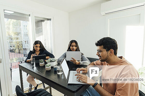 Lächelnder Mann mit Laptop vor Frauen  die über Tablet-PCs auf dem Tisch im Wohnzimmer lernen