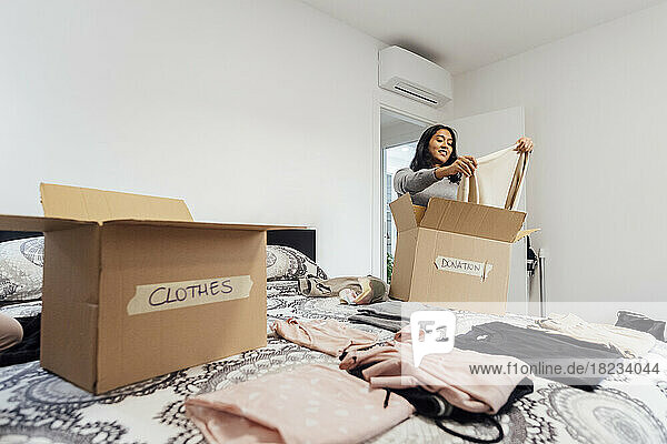 Smiling young woman packing box of clothes for donation on bed