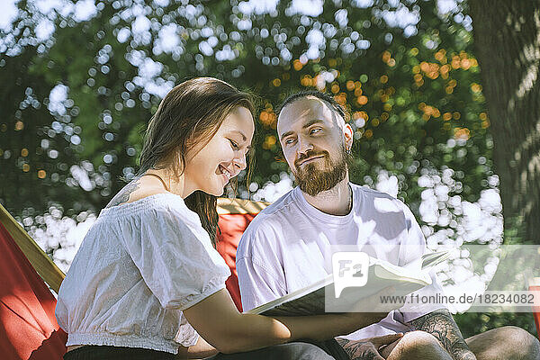 Hipster man looking at girlfriend smiling and reading book in backyard