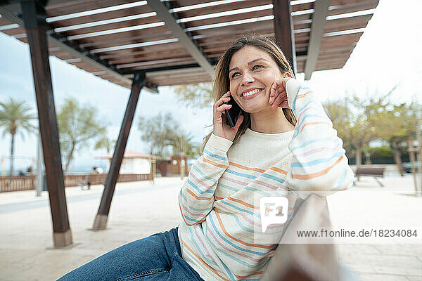 Smiling beautiful woman sitting on bench talking through mobile phone