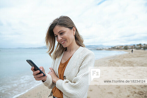 Lächelnde schöne junge Frau  die am Strand ihr Smartphone benutzt