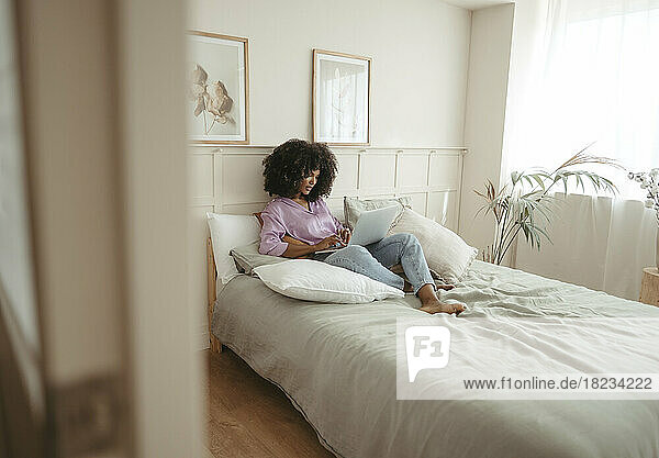 Young woman using laptop sitting on bed in bedroom