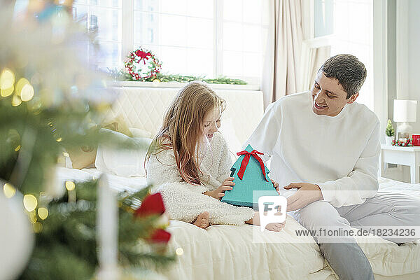 Happy girl holding Christmas gift with father sitting on bed at home