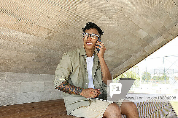 Smiling freelancer talking through mobile phone sitting with laptop on bench