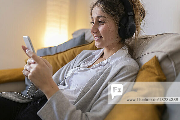 Smiling woman wearing wireless headphones using smart phone on sofa at home