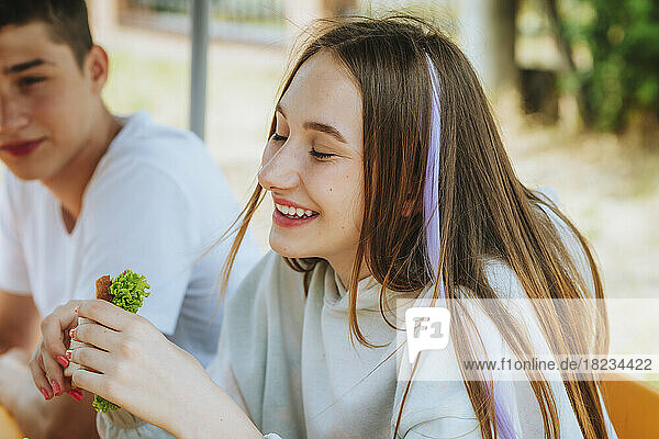 Happy teenage girl eating sandwich with friends in schoolyard