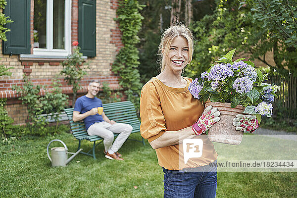 Smiling woman holding flowering plant with man in background at back yard