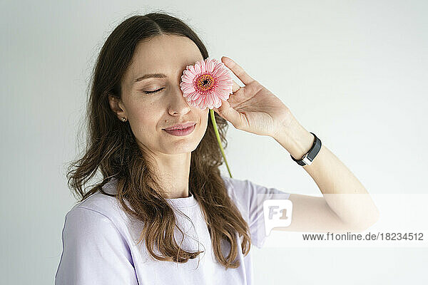 Smiling woman covering eye with gerbera flower in front of white wall