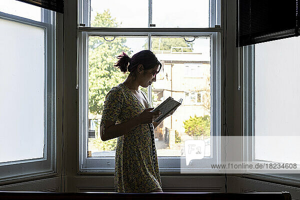 Smiling woman reading book standing in front of window