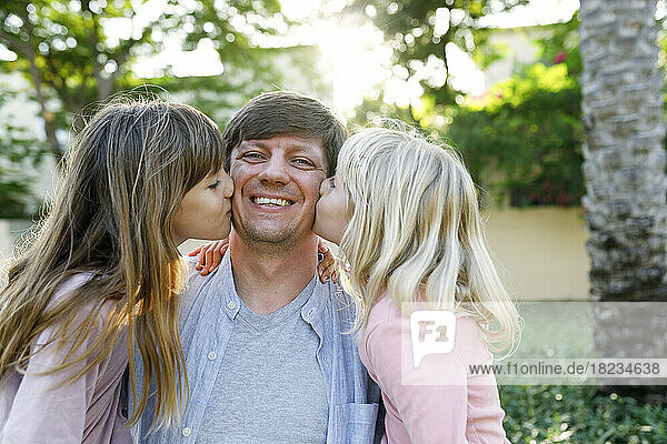 Happy father with daughters kissing him on cheeks at park