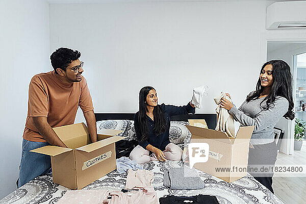 Glückliche Frauen und Männer packen zu Hause Kisten mit Kleidung für eine Spende auf dem Bett