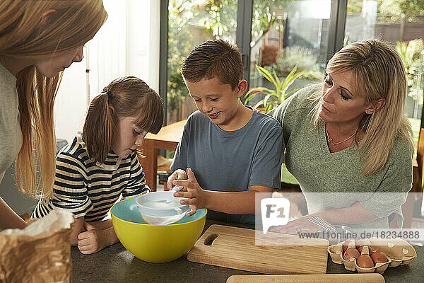 Mutter und Schwestern schauen einem Jungen zu  der zu Hause Mehl siebt