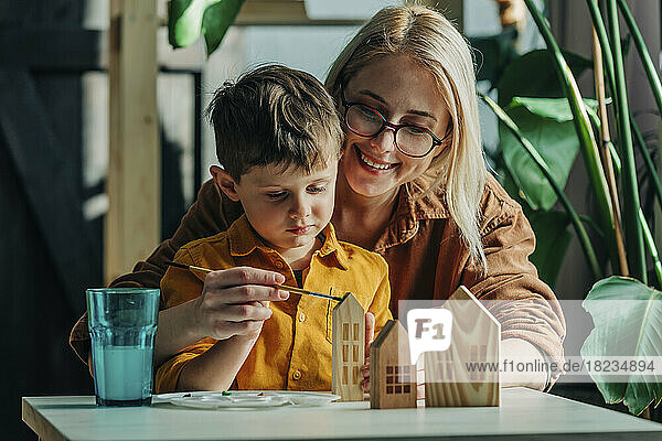 Happy mother and son painting wooden model houses at table