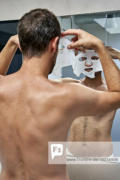 Man applying sheet mask on face in front of mirror at home