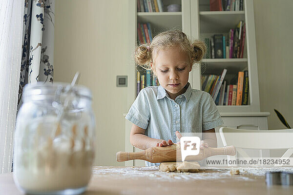 Blond girl rolling cookie dough on table at home