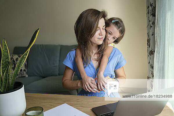 Tochter umarmt Mutter  die mit Laptop am Schreibtisch im Heimbüro sitzt