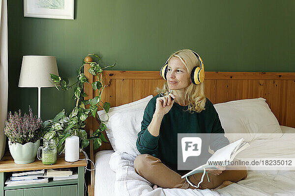 Contemplative woman wearing headphones with notebook in bedroom