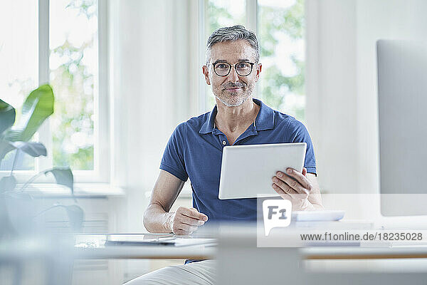 Smiling mature doctor sitting with tablet PC at desk