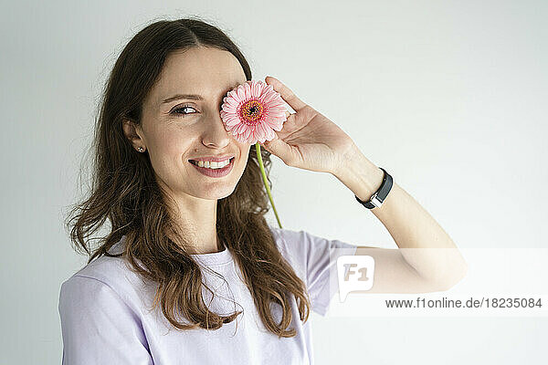 Happy woman covering eye with gerbera flower in front of white wall