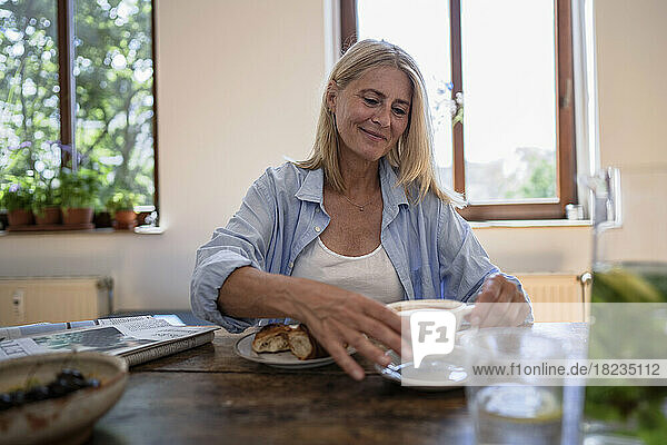 Smiling woman picking up coffee cup sitting at table in home