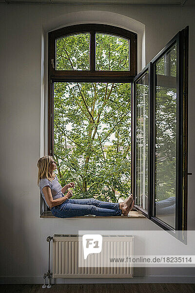 Contemplative mature woman sitting on window sill at home