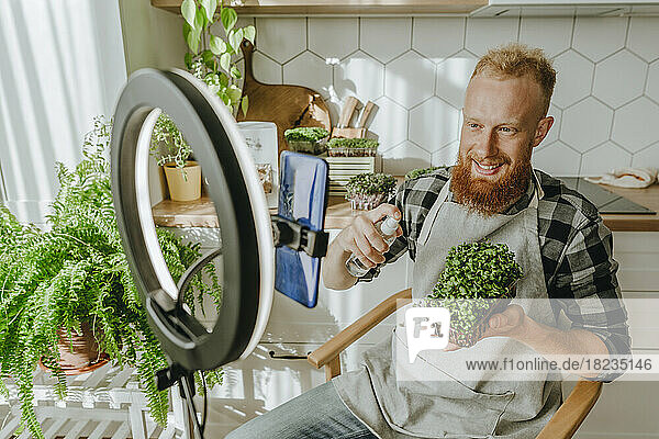 Smiling man spraying water on microgreens sitting in front of smart phone