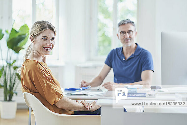 Happy woman sitting on chair with doctor in background at medical practice