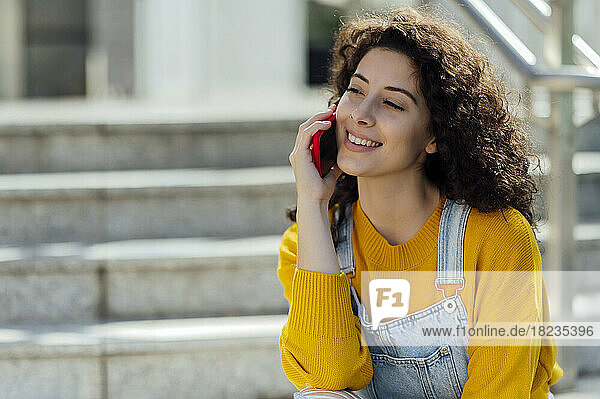 Lächelnde junge Frau mit lockigem Haar  die auf der Treppe über ihr Mobiltelefon spricht