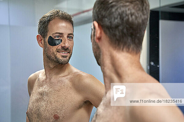 Happy man looking in mirror with eye patch on face at home