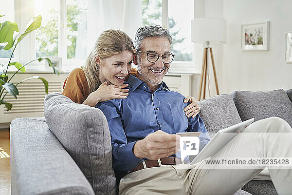 Happy woman with arm around man with credit card online shopping through table PC on sofa